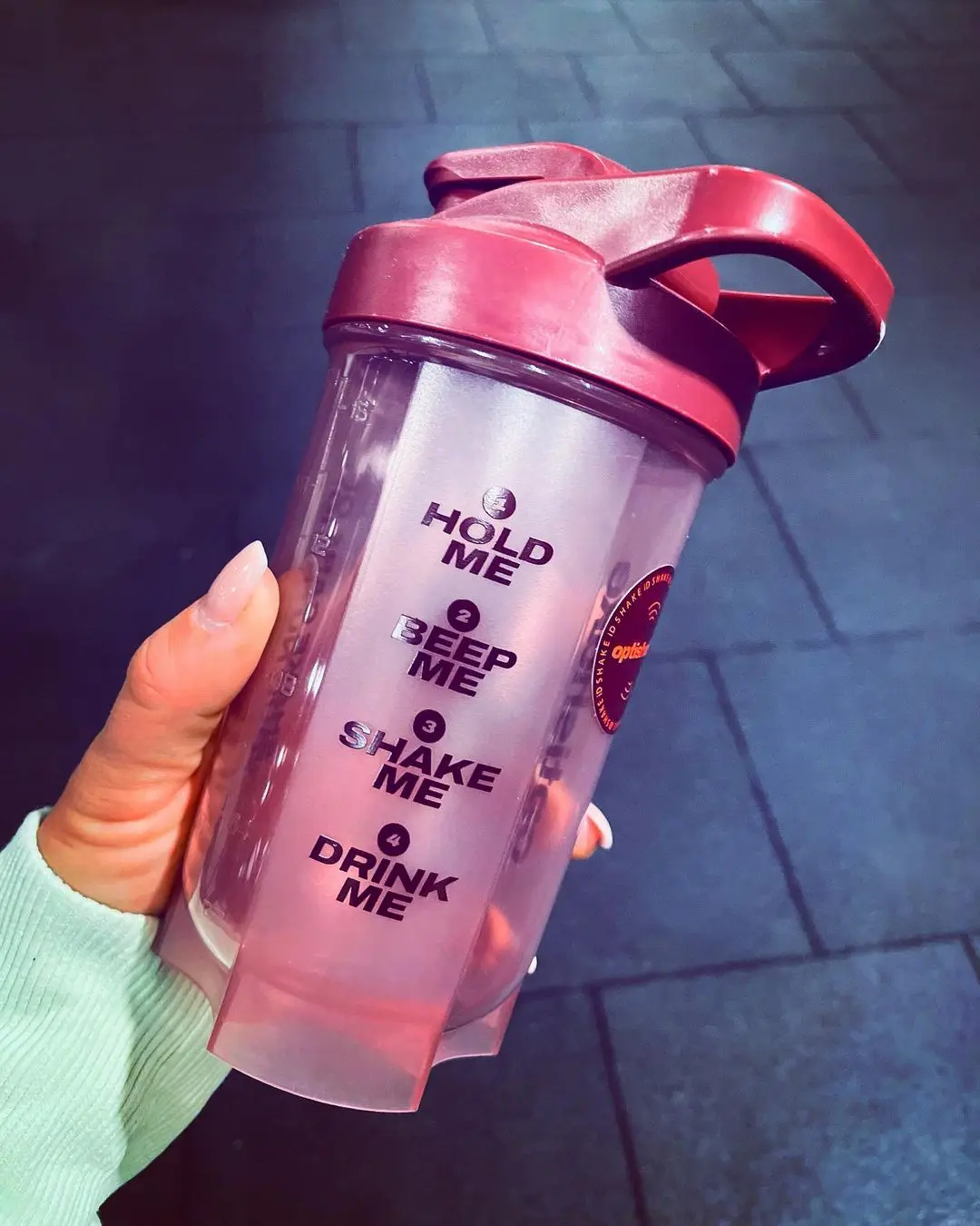 An Optishake shaker held in hand, printed with its four-step flow: hold, beep, shake, drink
